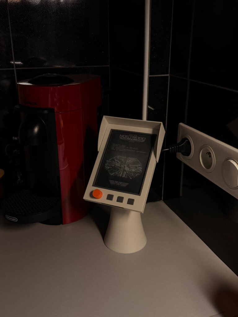 Kindle dans le support imprimé en 3D de QALC, posée dans la cuisine à côté d'une machine à café, molette orange et trois touches