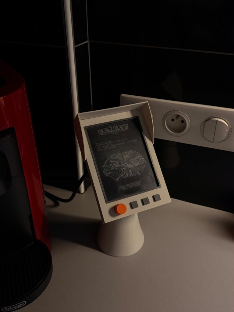 Gros plan du support Nostrum : écran e-ink USCSS Nostromo, visière, molette orange et trois touches grises